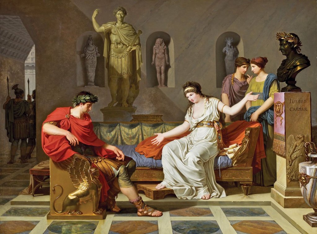 A historical scene depicting Julius Caesar seated in a Roman interior, engaged in conversation with a woman dressed in white, while three other women observe. Statues and a bust of Caesar are visible in the background.